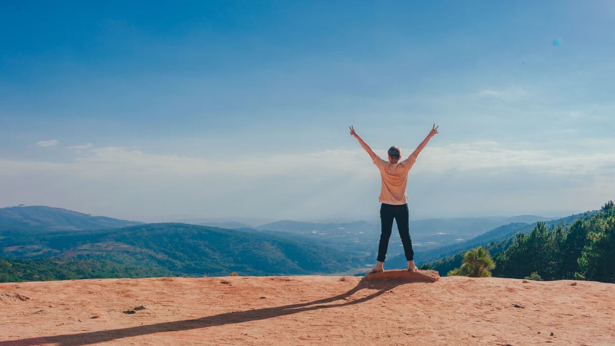 A man raises his arms in triumph on a rocky mountain summit overlooking a vast landscape